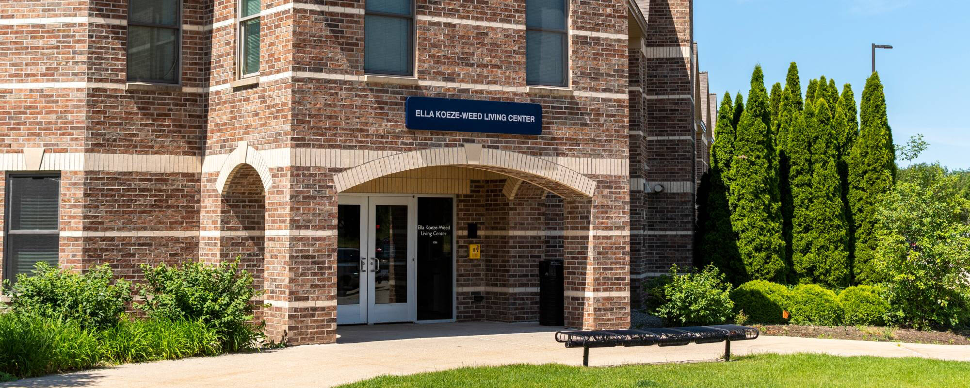 Brick building with "Ella Koeze-Weed Living Center" sign, arched entryway, and surrounding greenery on a sunny day, conveying a welcoming tone.